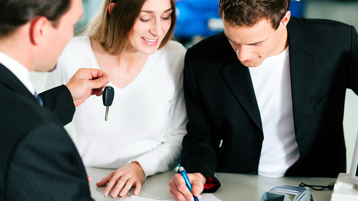 Woman smiling as man signs paper while the dealership employee hands the key to them